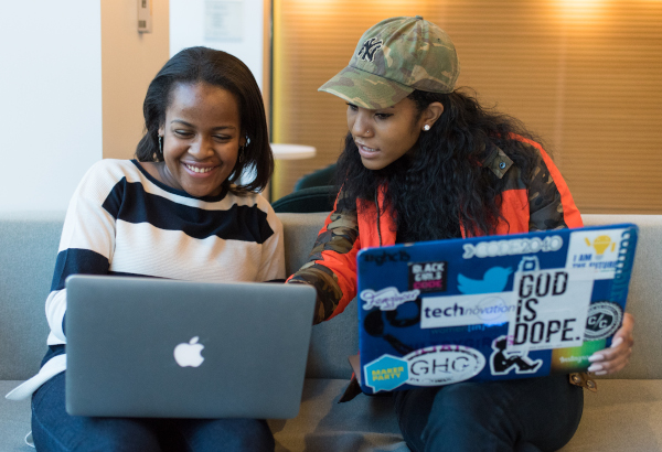 Two black women sit on a couch, with laptops on their lap. One woman is pointing to the other woman's laptop screen. The other woman is smiling. Photo is cc-by 2.0 WOCinTech Chat